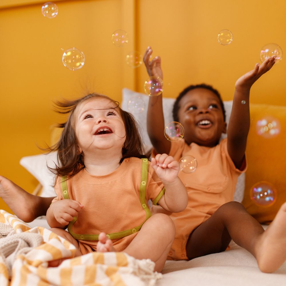 Two young children sat on a bed wearing orange outfits and reaching up to catch bubbles 