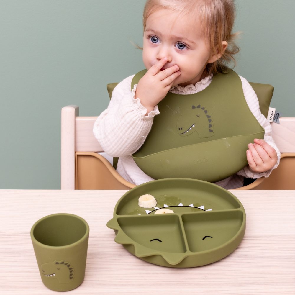 A child in a highchair wearing a dinosaur themed bib with a dinosaur themed plate and cup 
