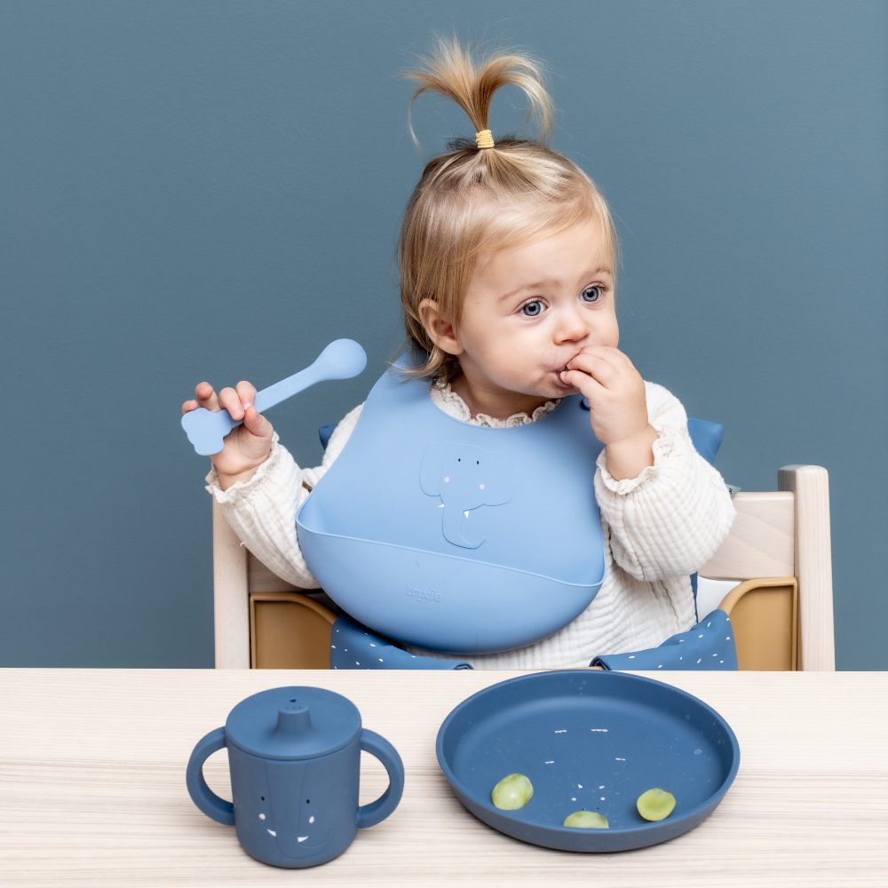 A child in a blue elephant bib with a blue cup and bowl from the Trixie tableware range 
