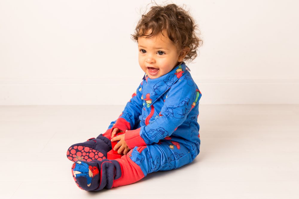 A baby sat on the floor wearing a Paddington Bear romper and matching bib and booties 