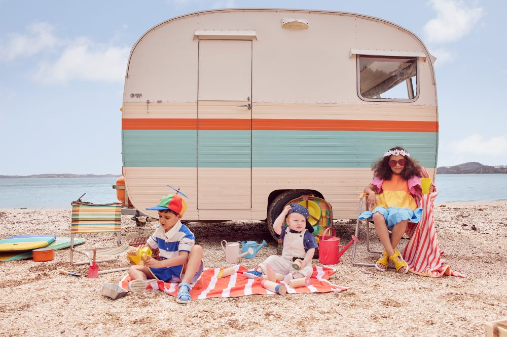 Children on a beach in front of a vintage-style caravan