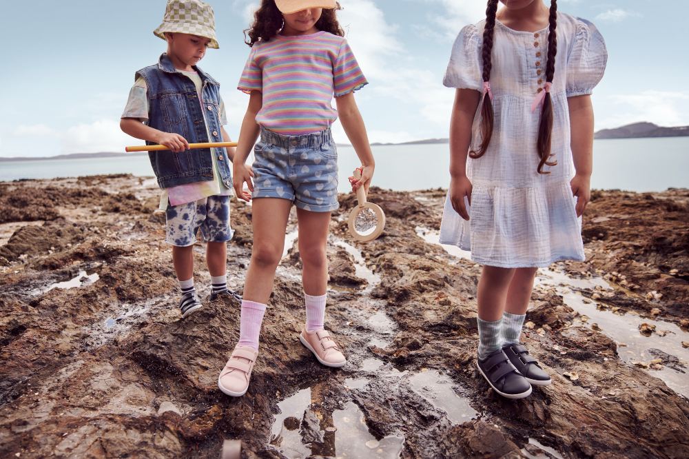 Three children walking across rocks on a beach wearing Bobux shoes 