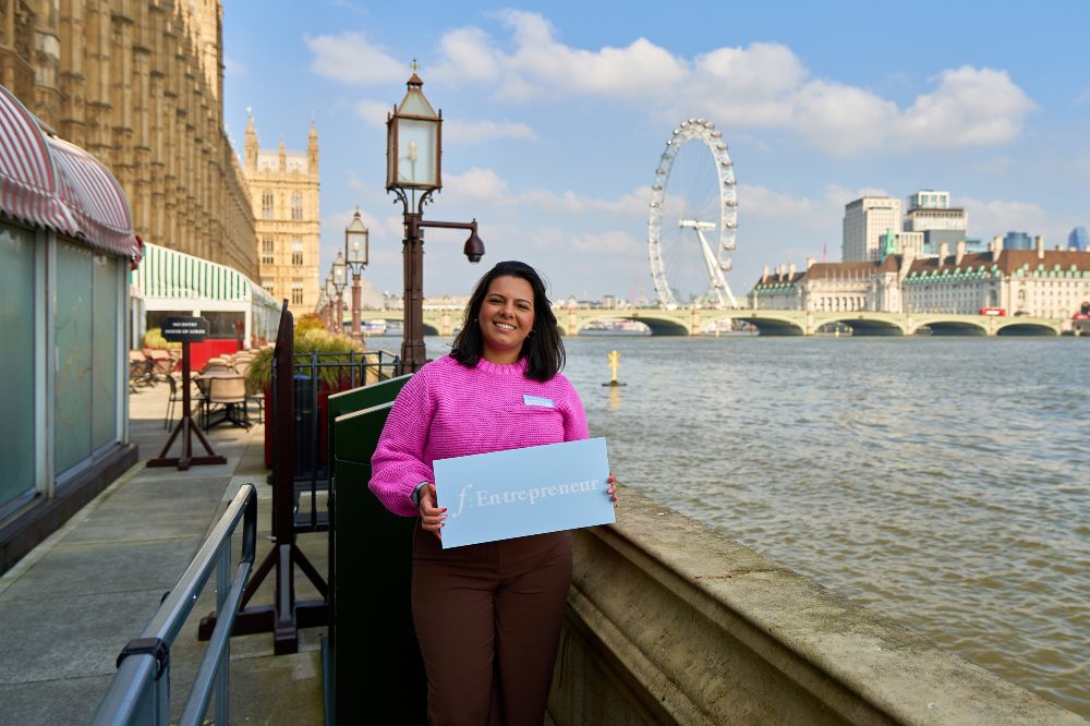 A woman stood beside the Thames holding up a board saying F:Entrepreneur as part of a celebration of female entrepreneurs