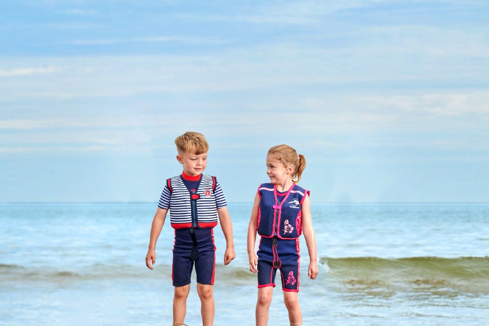 A young boy and girl stood by the sea wearing swim jackets by Konfidence