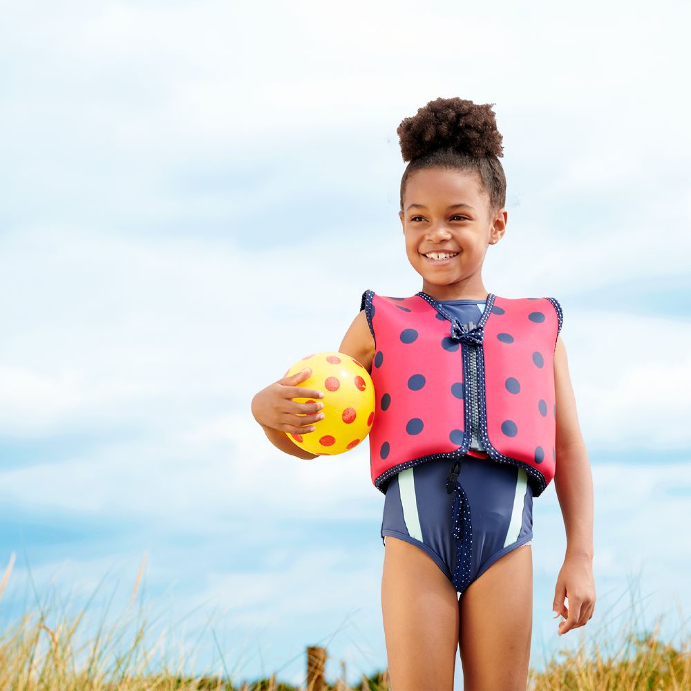 A young girl stood outside holding a ball and wearing a red swim jacket with blue spots by Konfidence