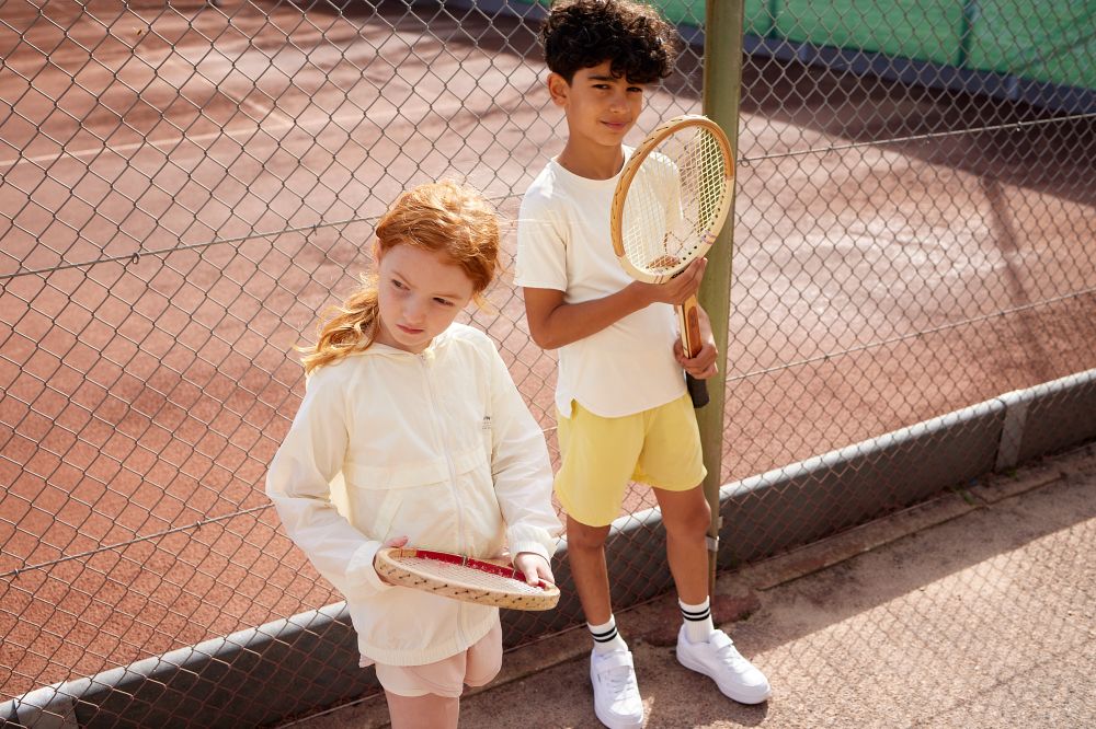 A young girl and boy stood on a tennis court holding rackets and wearing shorts and tops from the MINI A TURE Activewear collection