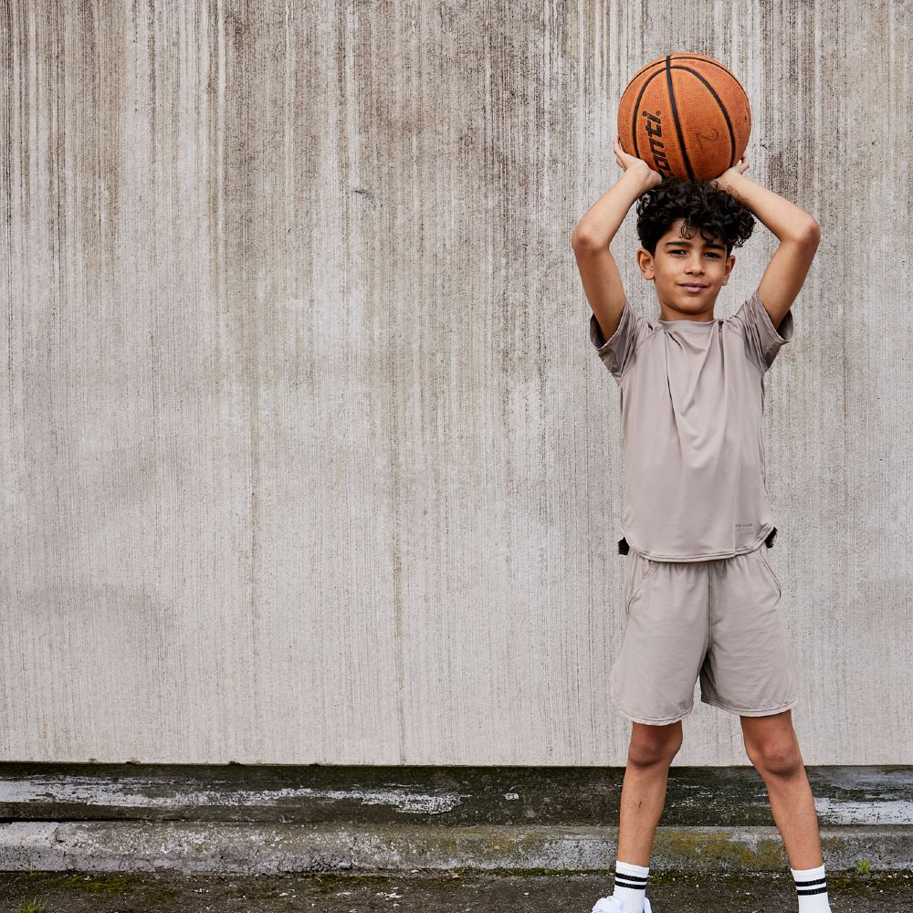 A young boy stood in front of a wall wearing shorts and T-Shirt and holding ball above his head