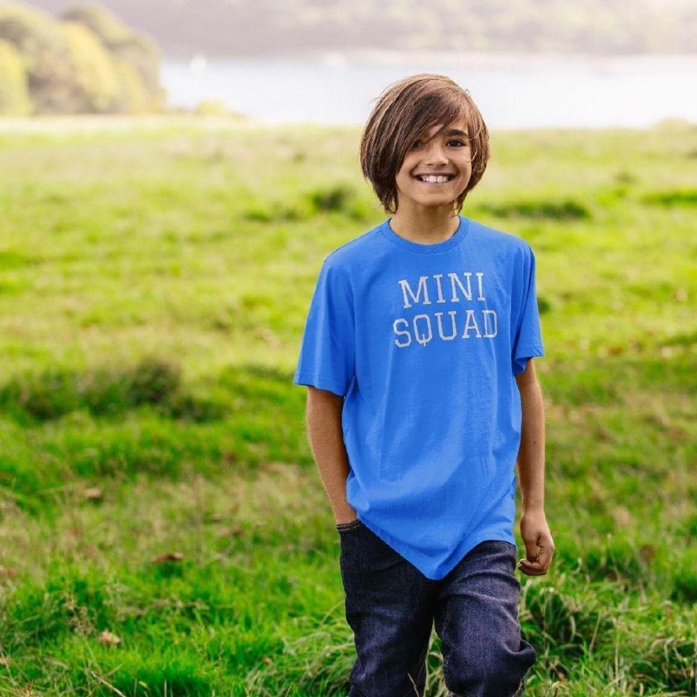 A young boy stood outside wearing jeans and a blue T-shirt with Mini Squad written on the front