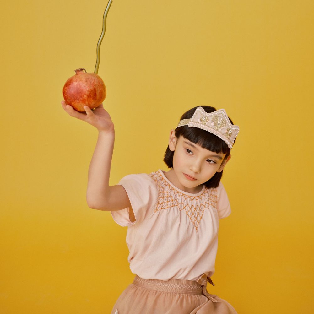A young girl wearing a pink crown headdress holding up a pomegranate with a metal straw in it 