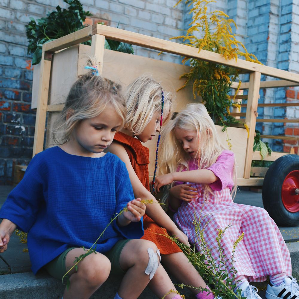 Three children sat outside in front of a wooden cart playing with plants wearing bright outfits by childrenswear brand Cacane 