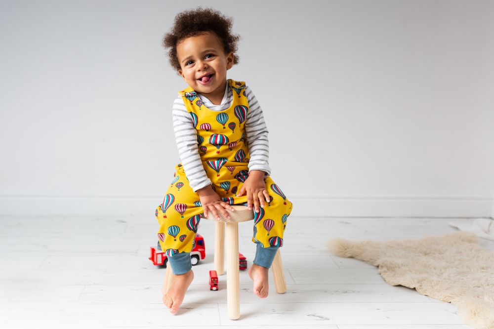 A child sat on a stool wearing yellow dungarees with a hot air balloon print 