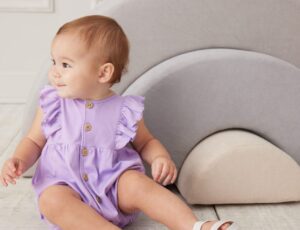 A young baby sat down wearing a lilac romper with frills and buttons down the front