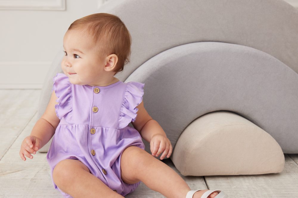 A young baby sat down wearing a lilac romper with frills and buttons down the front