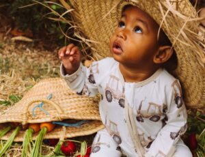 A young baby sat outside wearing a straw hat and tractor print babygro