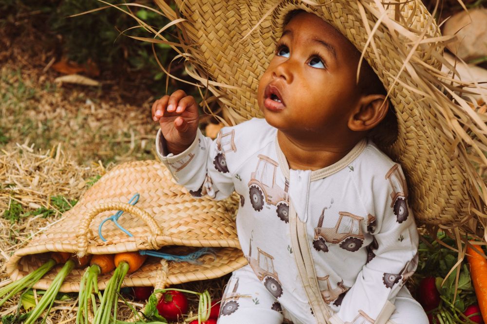 A young baby sat outside wearing a straw hat and tractor print babygro