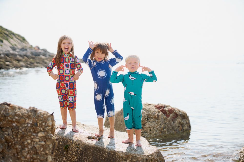 Three children stood on a rock in the sea wearing swimwear by Muddy Puddles
