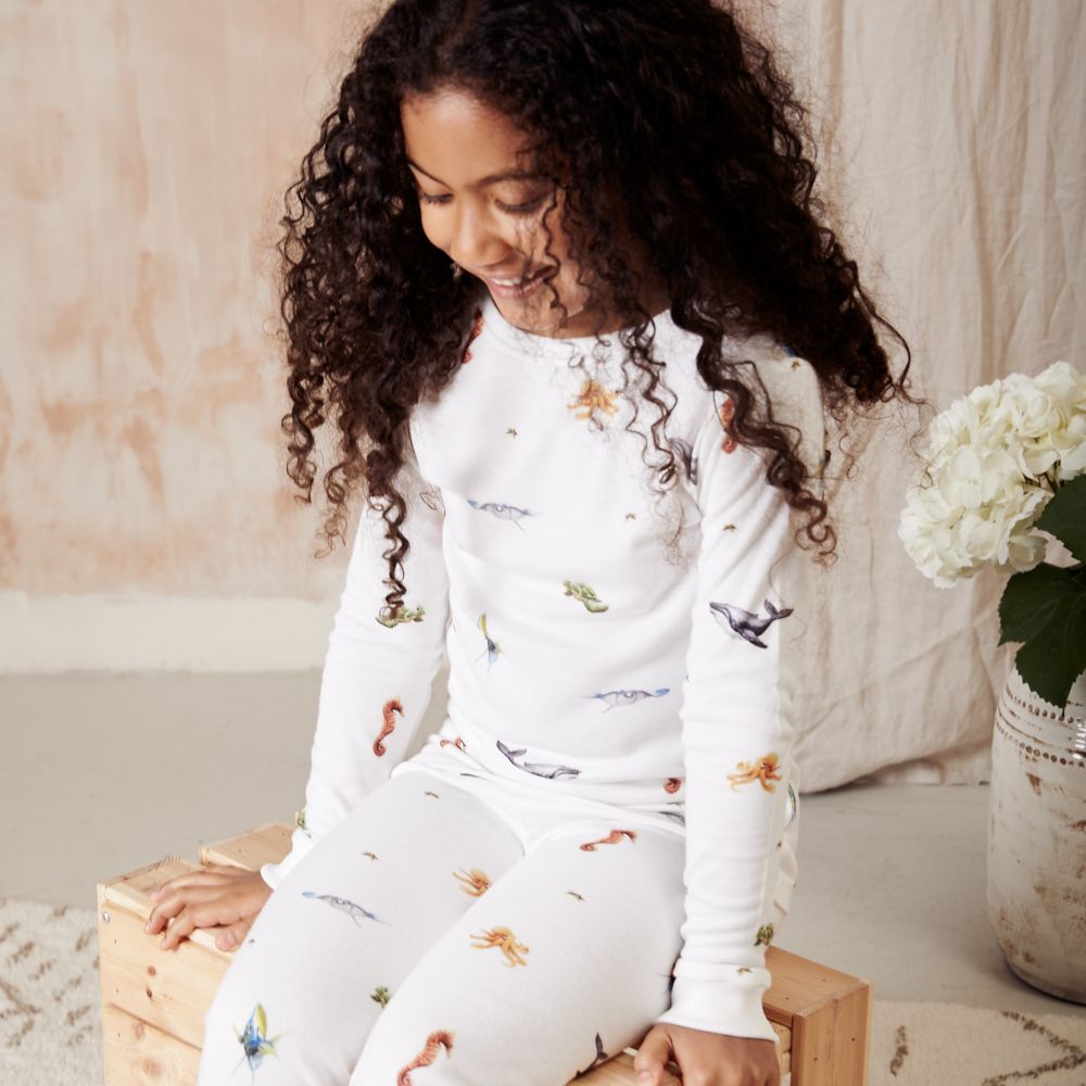 A girl with long, dark curly hair sat on a wooden box wearing pyjamas by Rosa & Blue 