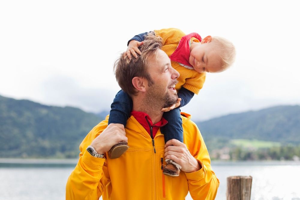 A man stood in front of a lake in a yellow coat with a young child on his shoulders in a matching yellow coat