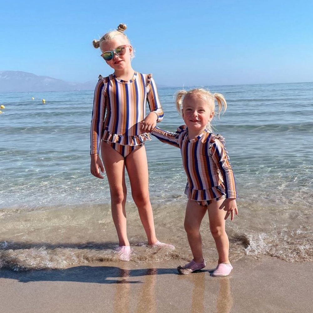 Two young girls in swimwear and beach shoes stood on a beach 