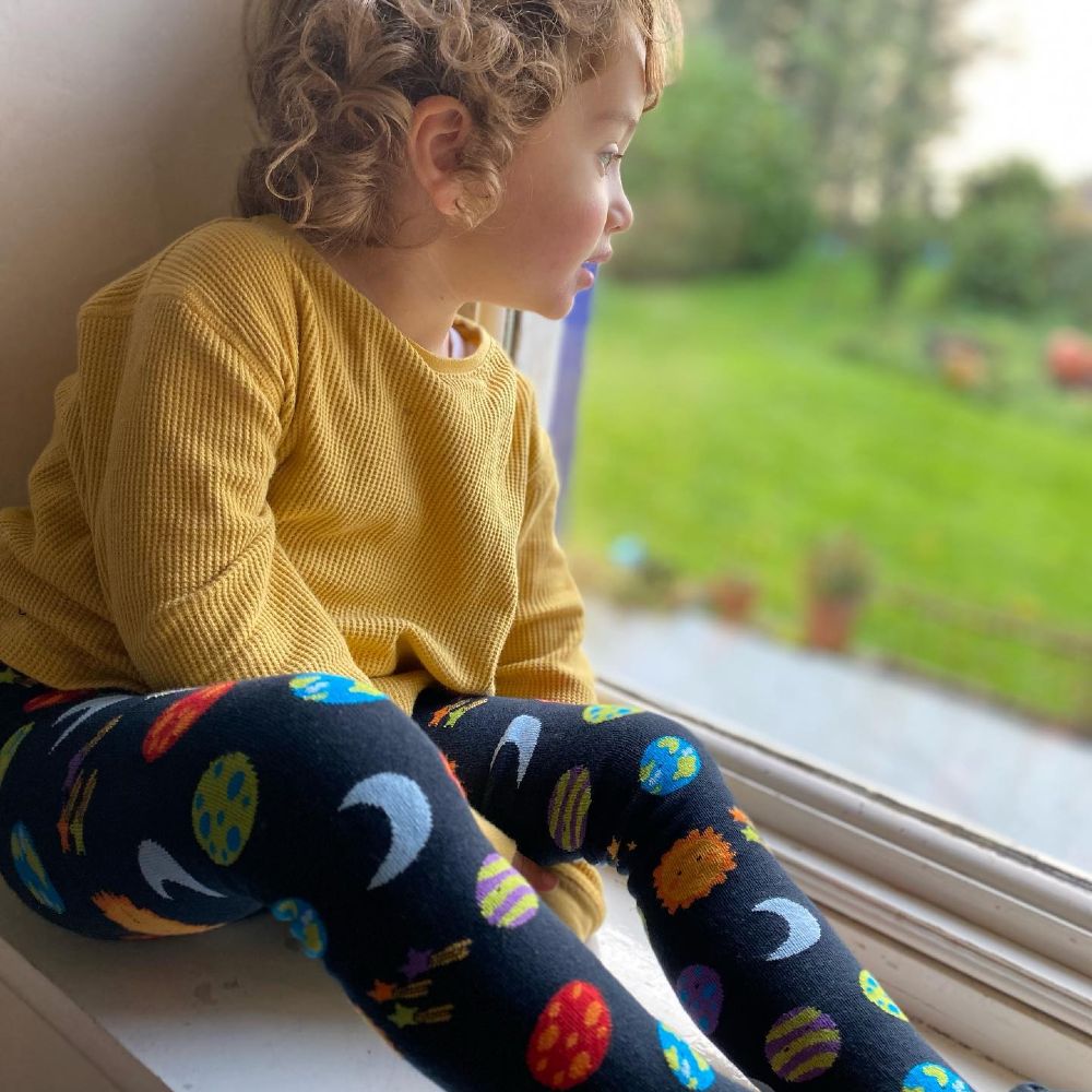 A child sat in a window sill wearing a yellow top and footless tights with planets on them
