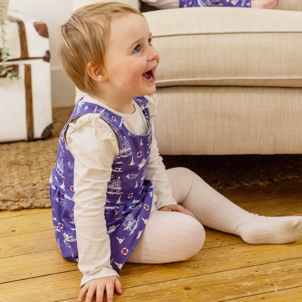 A toddler sat on the floor beside a sofa wearing a blue dress and white tights 