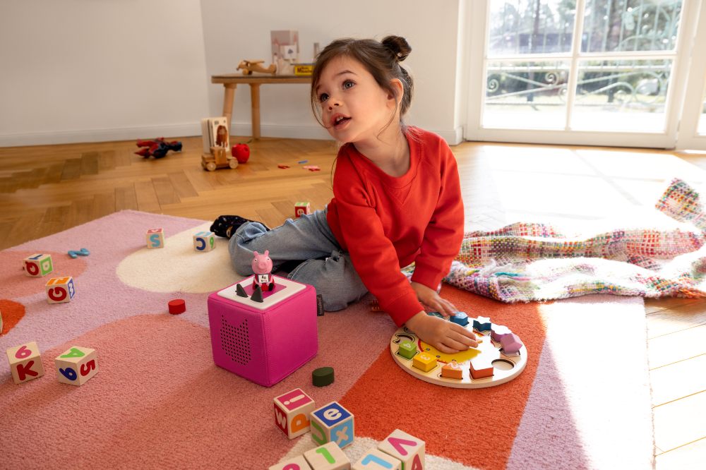 A child sat on the floor playing with toys in front of a pink Leann with Peppa Toniebox