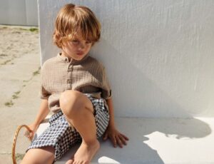 A young boy sat on the fall against a white wall wearing shorts and T-shirt by Wheat Clothing