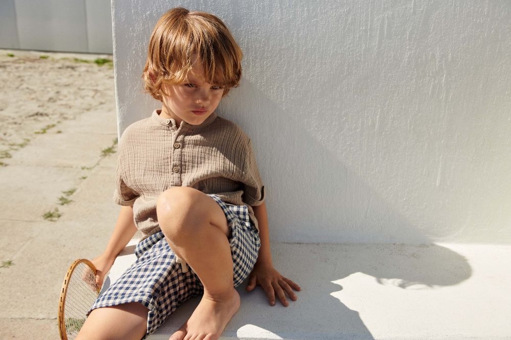 A young boy sat on the fall against a white wall wearing shorts and T-shirt by Wheat Clothing