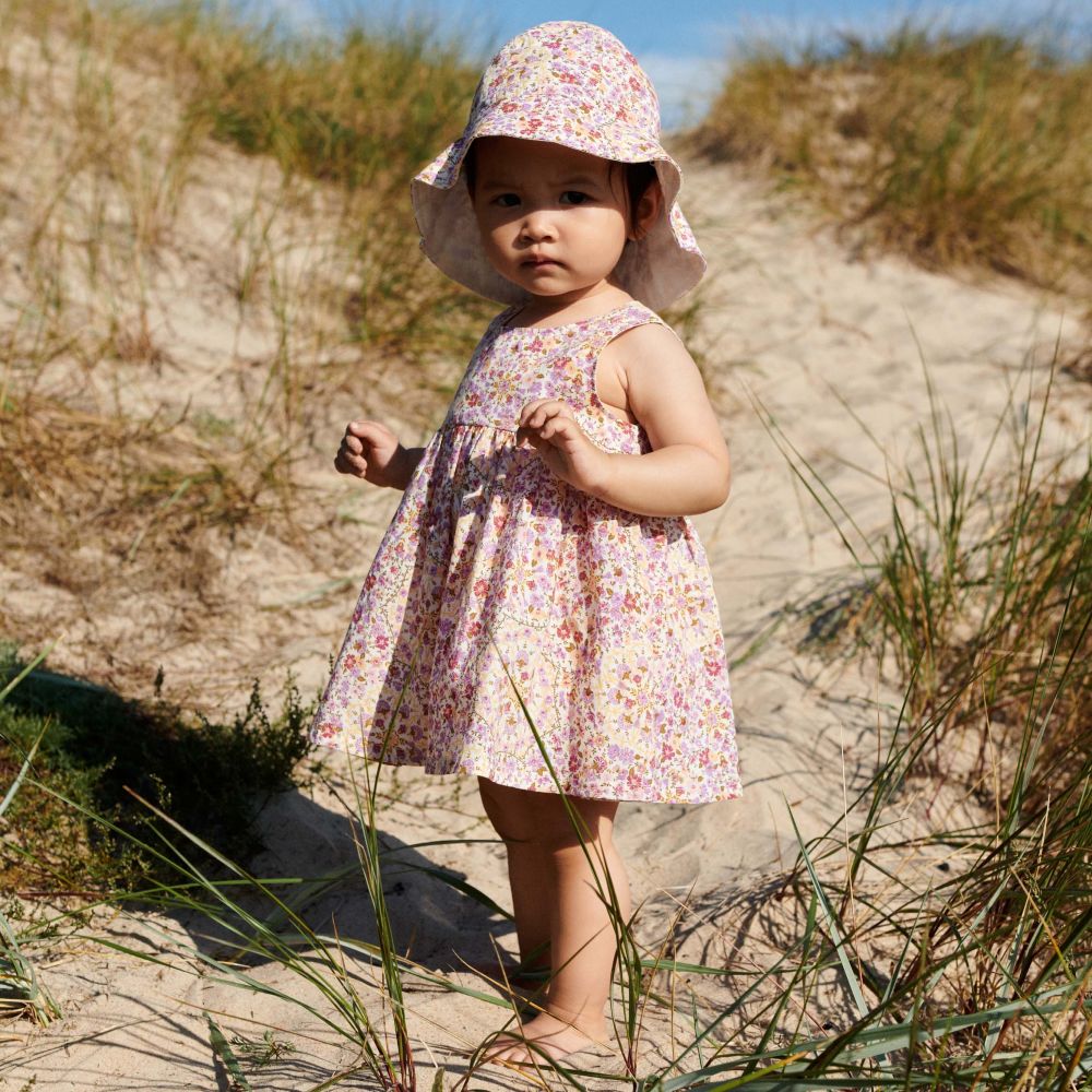 A young girl stood on a beach in sand dunes wearing a sun dress and hat 