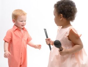 A young girl in a pink dress holding a microphone beside a young boy in an orange boilersuit