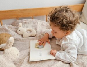 A young child lying on a bed reading a book beside a rabbit teddy and rattle