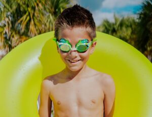 A young boy with an inflatable rubber ring around his shoulders wearing green serpent swim goggles by Bling2o