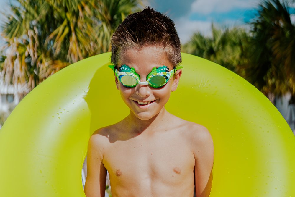 A young boy with an inflatable rubber ring around his shoulders wearing green serpent swim goggles by Bling2o