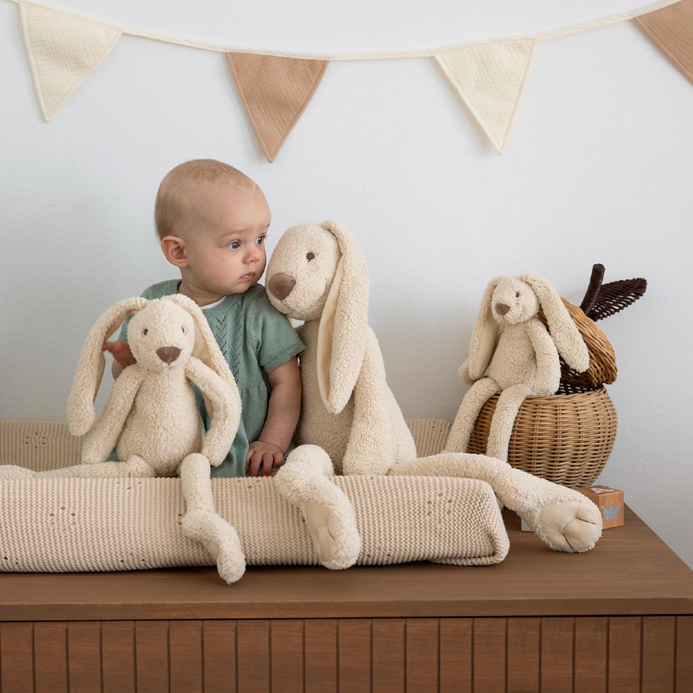 A baby sat on a changing table holding cream coloured rabbit soft toys 