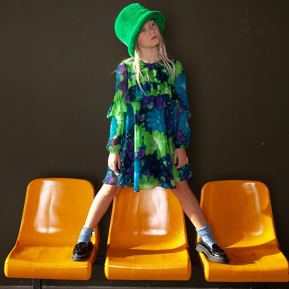 A girl stood on yellow plastic chairs wearing a green hat and a green and blue dress 