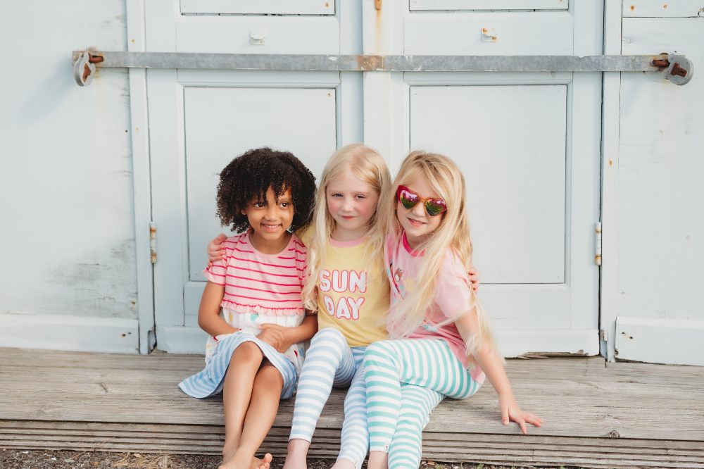 Three girls sat outside in front of wooden doors wearing outfits from the Happy Holidays collection by Lilly + Sid