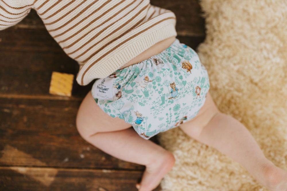 A baby crawling on a wooden floor wearing a striped jumper and a Peter Rabbit print cloth nappy by The Nappy Den