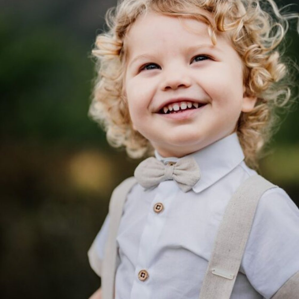 A young boy with blonde curly hair wearing a shirt, dicky bow and braces