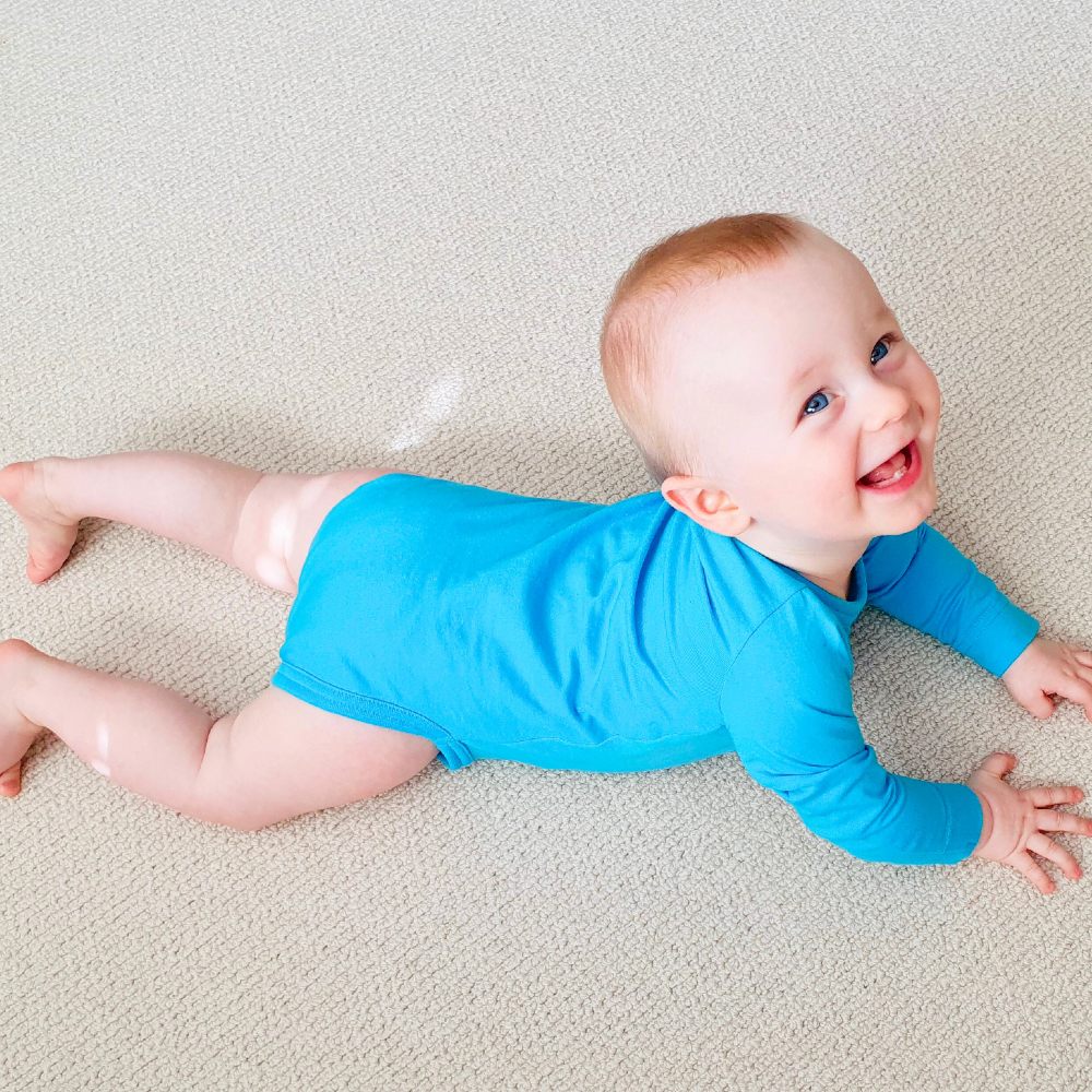 A baby lying on the floor wearing a bright blue babygro and laughing at the camera 