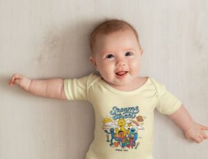 A baby lying down looking up at the camera wearing a yellow babygro with a Sesame Street print on the front