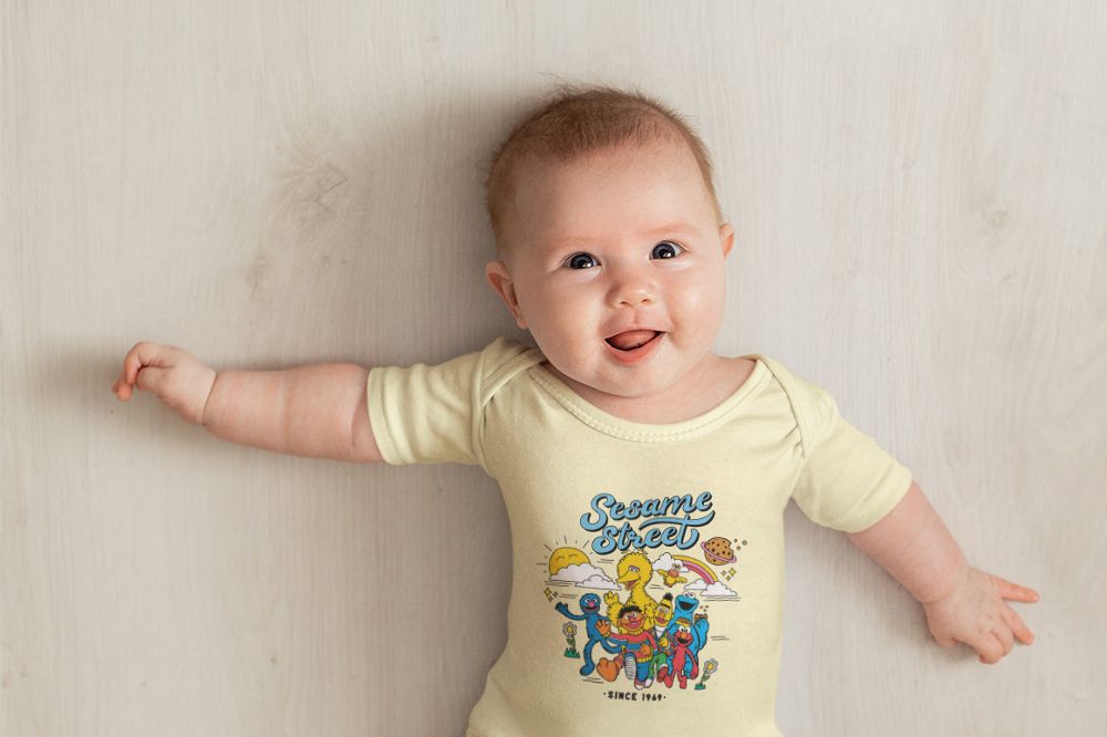 A baby lying down looking up at the camera wearing a yellow babygro with a Sesame Street print on the front