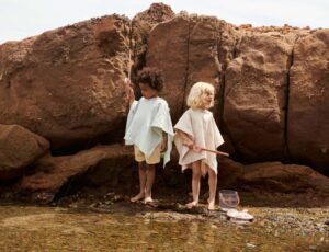 Two children stood on a shore by large rocks holding fishing nets and wearing towelling ponchos