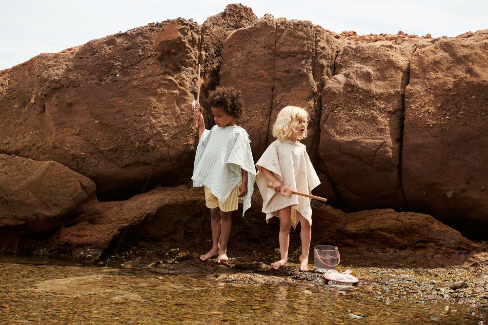 Two children stood on a shore by large rocks holding fishing nets and wearing towelling ponchos