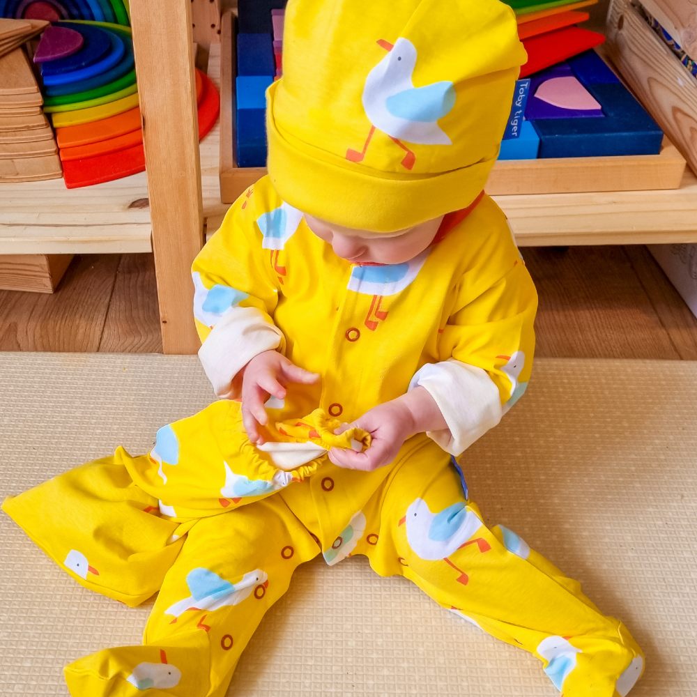 A young baby wearing a yellow babygro and hat with a seagull print on them 