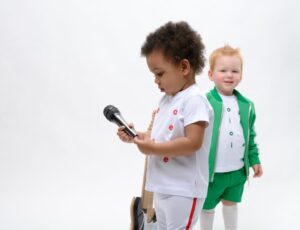 A a young child holding a microphone beside a young boy in a green jacket and shorts