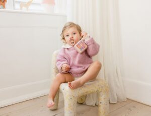 A young child in a pink jumper sat on a chair with a pacifier inn their mouth and a pacifier box in their hand from the BIBS x LIBERTY collection