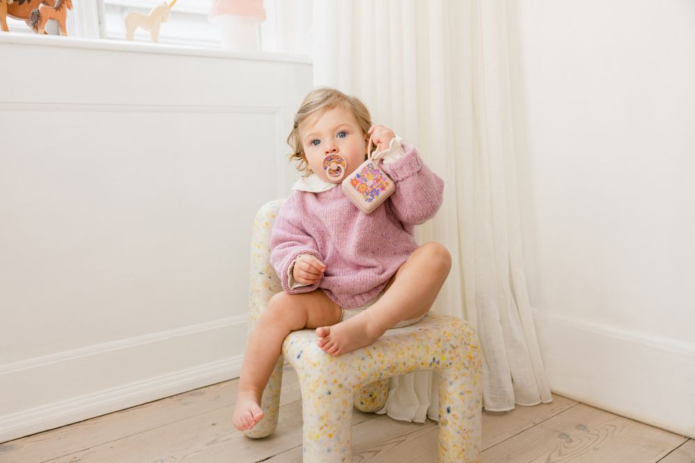 A young child in a pink jumper sat on a chair with a pacifier inn their mouth and a pacifier box in their hand from the BIBS x LIBERTY collection