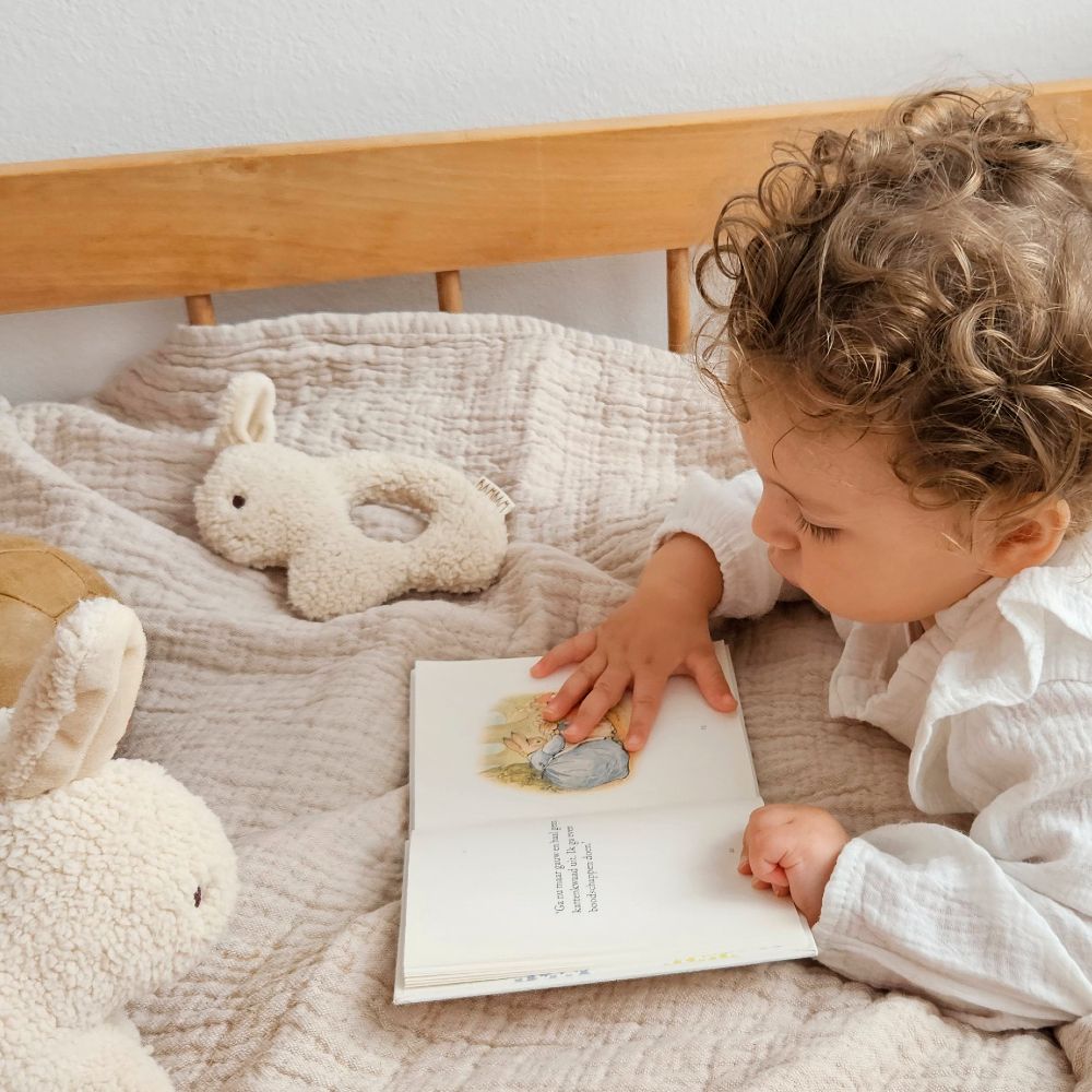 A toddler lying on a bed reading a book beside rabbit soft toys 