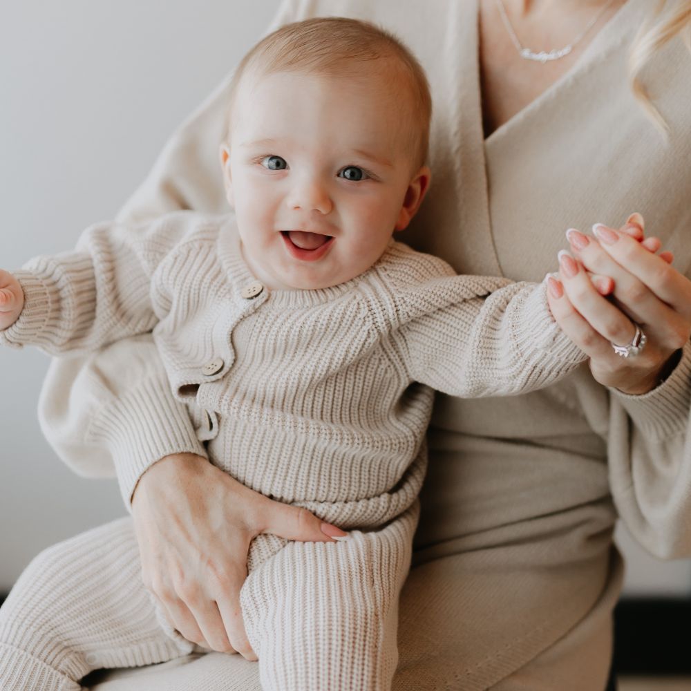 A young baby in a beige knitted top and trousers sat on someone's knee
