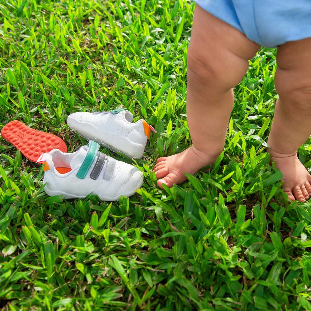 A toddlers legs show outside stood on grass beside a pair of shoes on the ground 